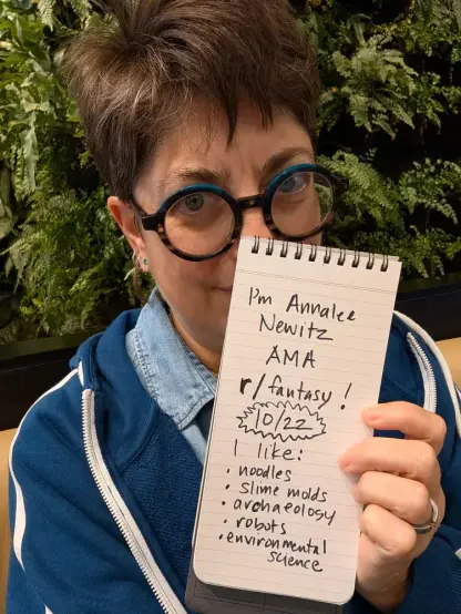 A picture of me in front of some plants. I'm holding a reporter's notebook open to a page that reads, in messy handwriting:

I'm Annalee Newitz 
AMA r/fantasy 10/22
I like:
Noodles
Slime molds 
Archaeology 
Robots
Environmental science