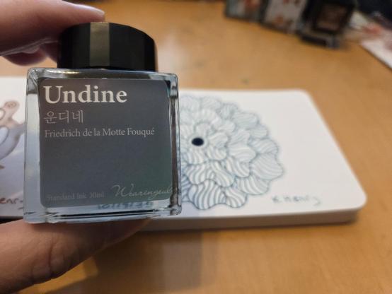 I'm holding a small bottle of ink, in one hand. Behind the bottle, is my daily ink drawing on an open page of my sketchbook. Today's pattern is floral, and its not finished yet in this photo.