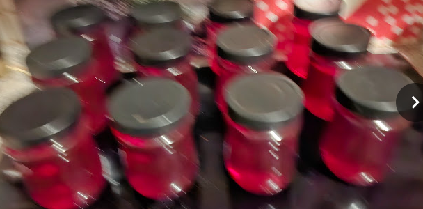12 glass jars containing orange-red quinces jelly arranged in rows on a dark glass surface. Each jar is topped with a black lid. The photo exhibits significant motion blur. The background contains indistinct red and white patterns. The jars occupy most of the frame.
