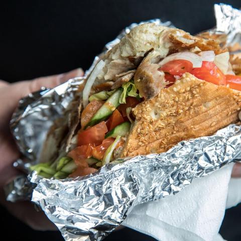 Nahaufnahme eines Döners im Sesamfladenbrot mit Seitan-Fleisch, Tomaten, Gurken und Salat, eingewickelt in Alufolie in einer Hand.