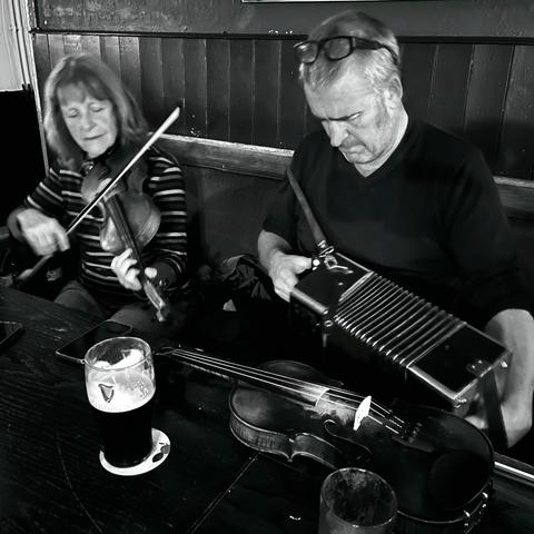 A woman playing fiddle and a man playing button accordion at a pub table.