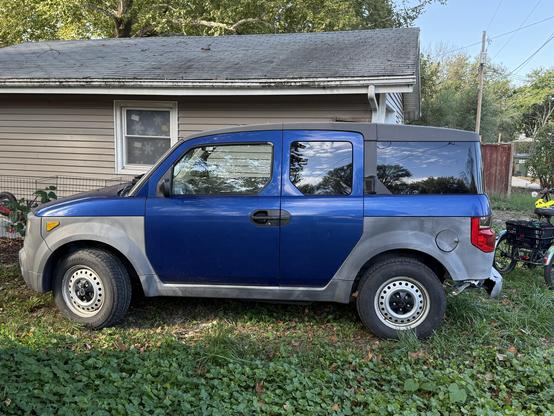 A Fiji blue Honda Element with worn gray quarter panels, steelie wheels, and part of the back left bumper missing sits in a backyard next to a  beige garage with a gray roof.