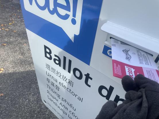 A large metal box with the words "Ballot Drop" partially obscured and in several different languages.  A red and white envelope is going in the narrow slot.