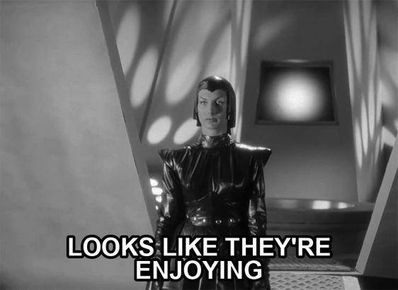 screenshot from the film Devil Girl From Mars showing a serious woman wearing a black leather suit, cape, and cowl. Text reads: "looks like they're enjoying"