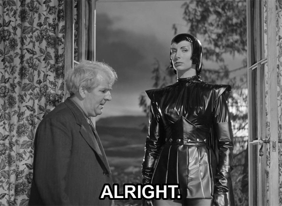 screenshot from the film Devil Girl From Mars showing a serious woman wearing a black leather suit, cape, and cowl. Text reads: "Alright."