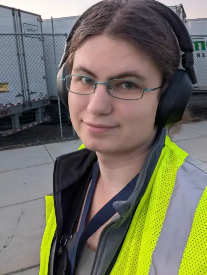 selfie of self in a hi-viz vest. trucks in the background. headphones on. walking home from work