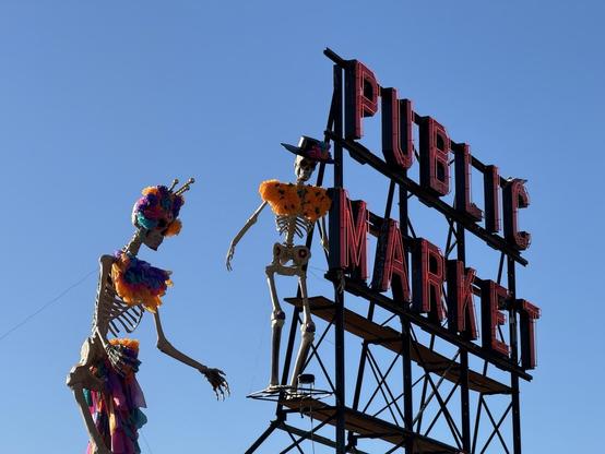 A colorful display featuring two skeleton figures adorned with floral decorations, positioned near a large "PUBLIC MARKET" sign against a clear blue sky.