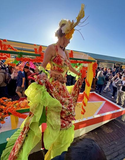 A person dressed in a vibrant floral costume and bright green fabric stands amidst a crowded festive atmosphere, with a backdrop of colorful decorations. The scene is illuminated by sunlight, creating a lively and celebratory mood.