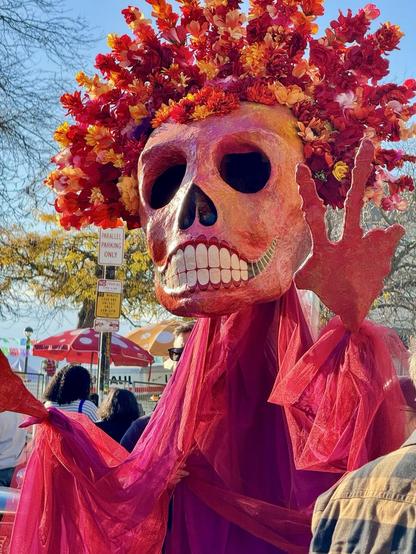 A large decorative skeleton figure adorned with vibrant flowers and a sheer red garment, standing in a festive outdoor setting. The skeleton is making a peace sign with its hand, surrounded by people and colorful umbrellas in the background.