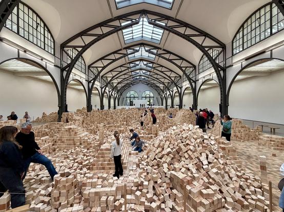 A huge hall with steel beams and white walls. It is filled with tons of wooden blocks. People of all ages are playing and building things