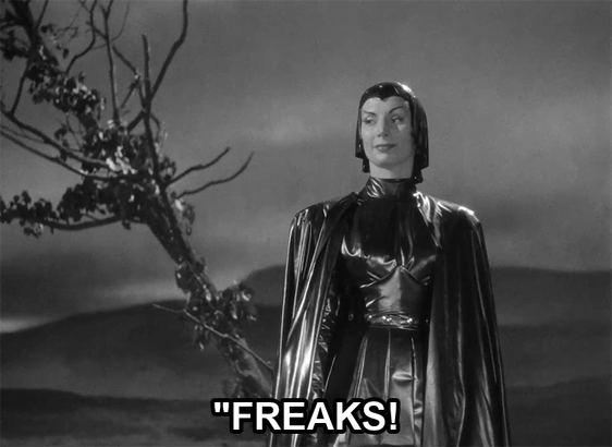 screenshot from the film Devil Girl From Mars showing a serious woman wearing a black leather suit, cape, and cowl. Text reads: ""Freaks!"