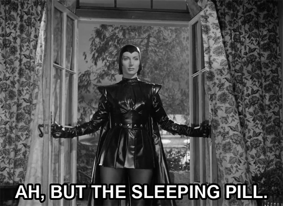 screenshot from the film Devil Girl From Mars showing a serious woman wearing a black leather suit, cape, and cowl. Text reads: "Ah, but the sleeping pill."