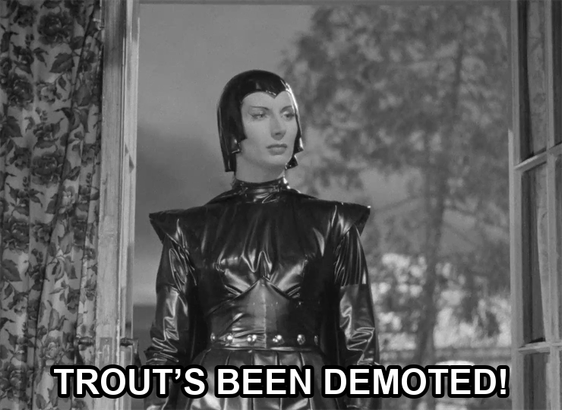 screenshot from the film Devil Girl From Mars showing a serious woman wearing a black leather suit, cape, and cowl. Text reads: "Trout’s been demoted!"