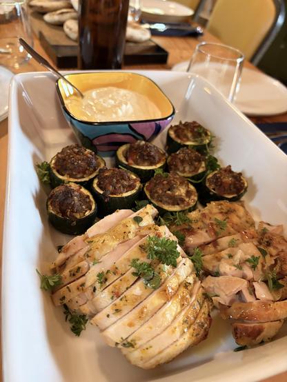 A plater with sliced chicken and stuffed zucchini. A square bowl of garlic yoghurt on the side.