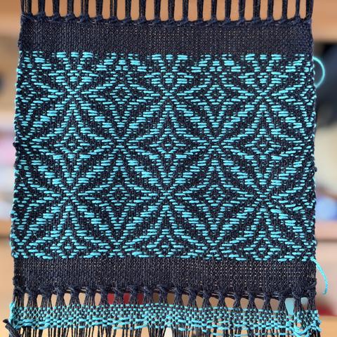 A black woven coaster on the loom has a teal overshot star pattern woven into it. With one mistake toward the end when my partner came out to talk at me. The black plain woven edges have been neatly hemstitched.